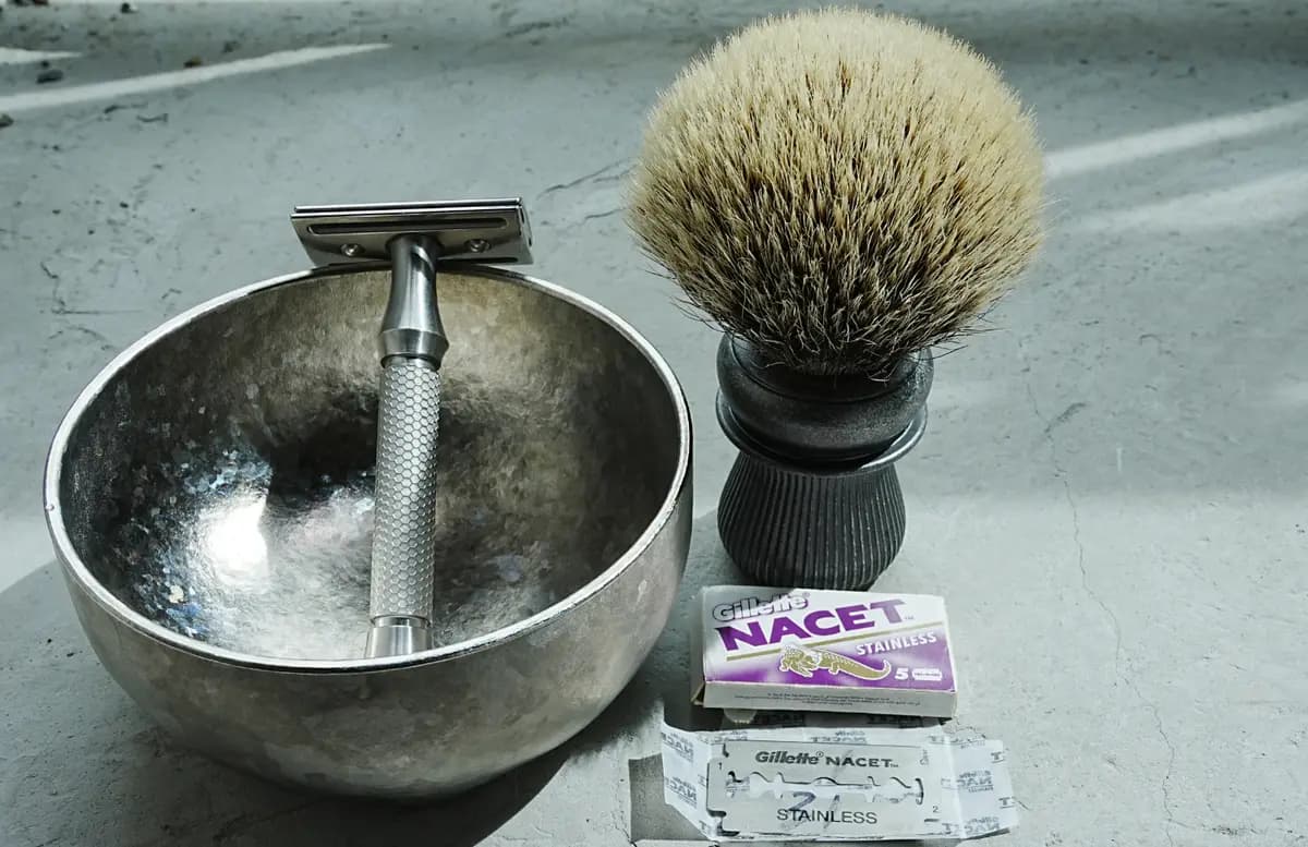 Gillette Nacet Stainless DE blade pack and a loose blade beside the ORMIXA Vector 316L safety razor, a titanium bowl and a badger brush on a concrete surface