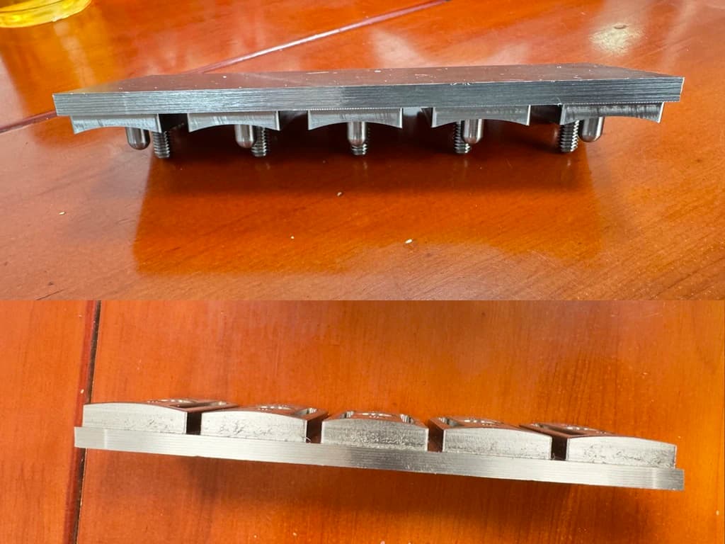 Side-profile photo of five Vector razor heads still attached to the billet stock, freshly machined and waiting to be flipped for the second-side machining pass — showing the original bar-stock thickness above and the cut head profile below