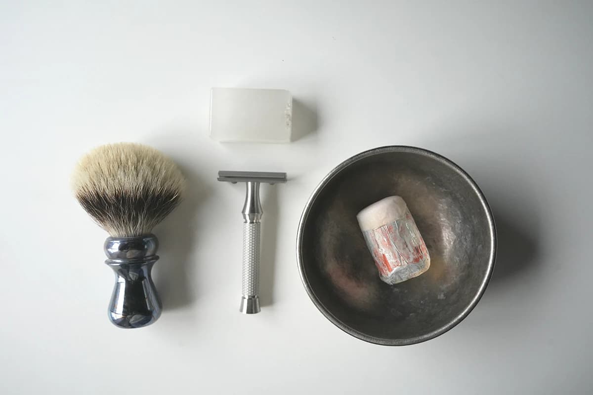 Top-down photo of the wet shaving testing kit on a neutral surface: shaving brush, ORMIXA Vector safety razor, alum block, and a soap puck in a metal lather bowl